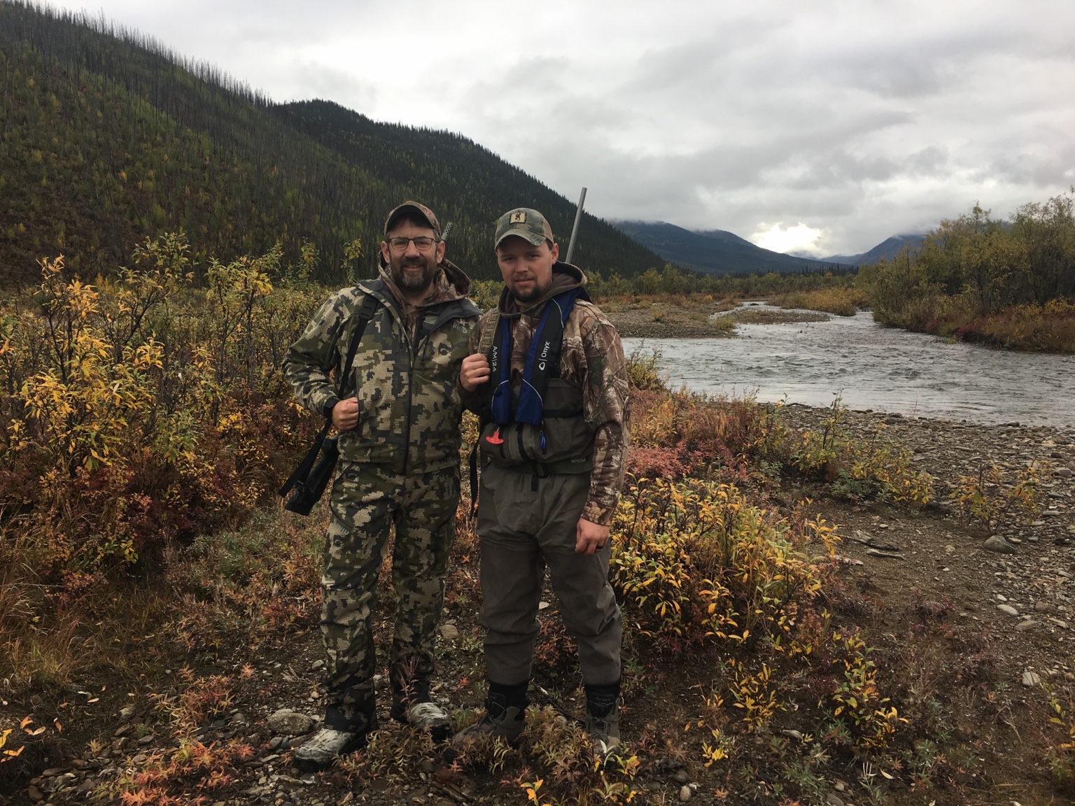 Tom Anderson with a hunting partner on a rocky Alaskan riverbank beneath forested mountains